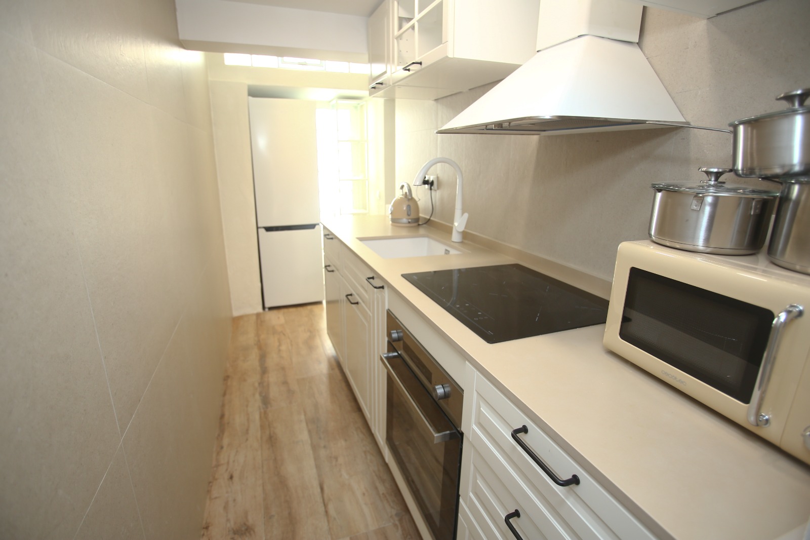 A narrow finished kitchen: white cabinets, cream counter, black hob, daylight at the far end.