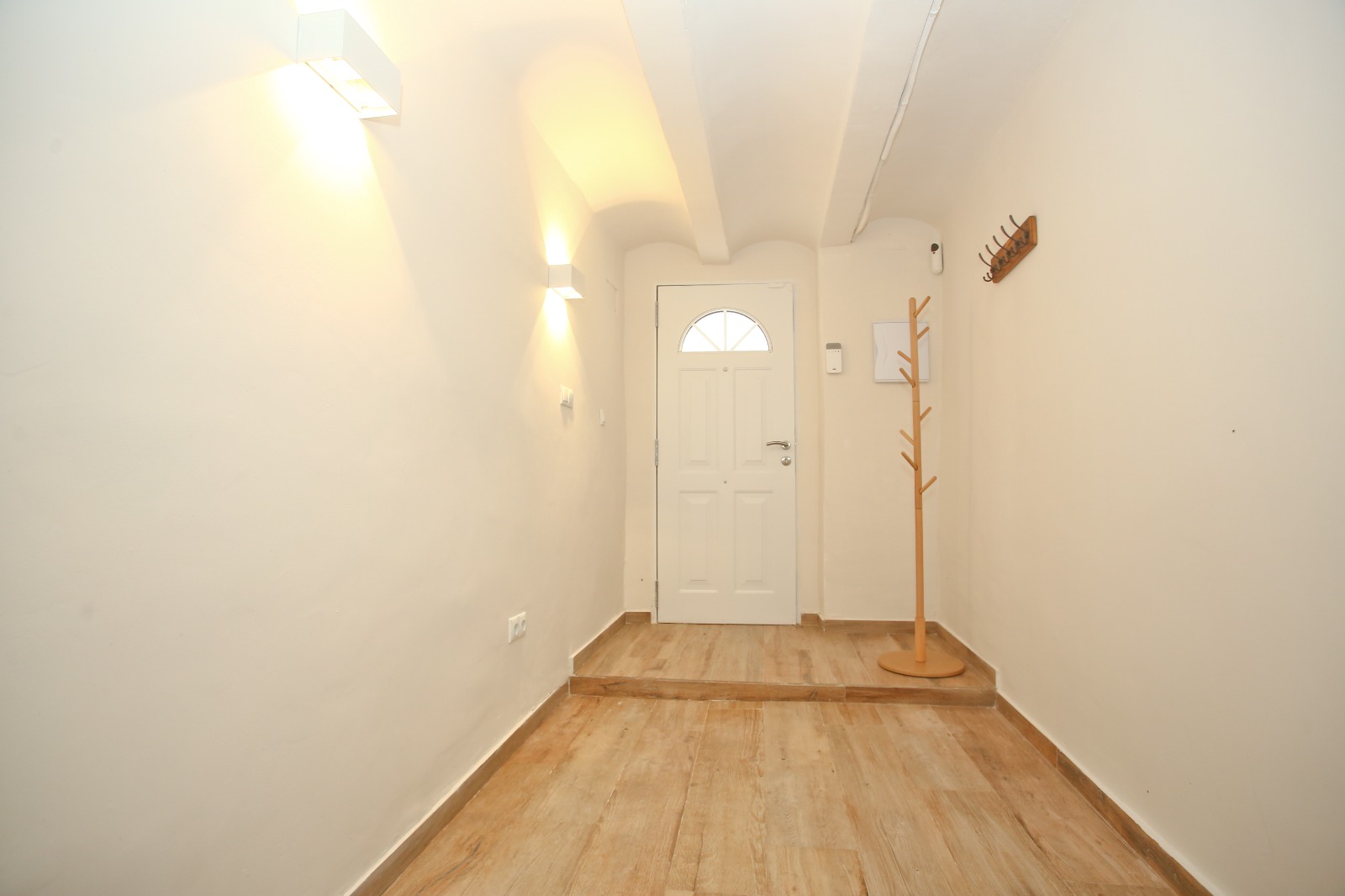 The finished entry: vaulted white ceiling, oak-look floor, wall sconces, a white door with a fanlight.