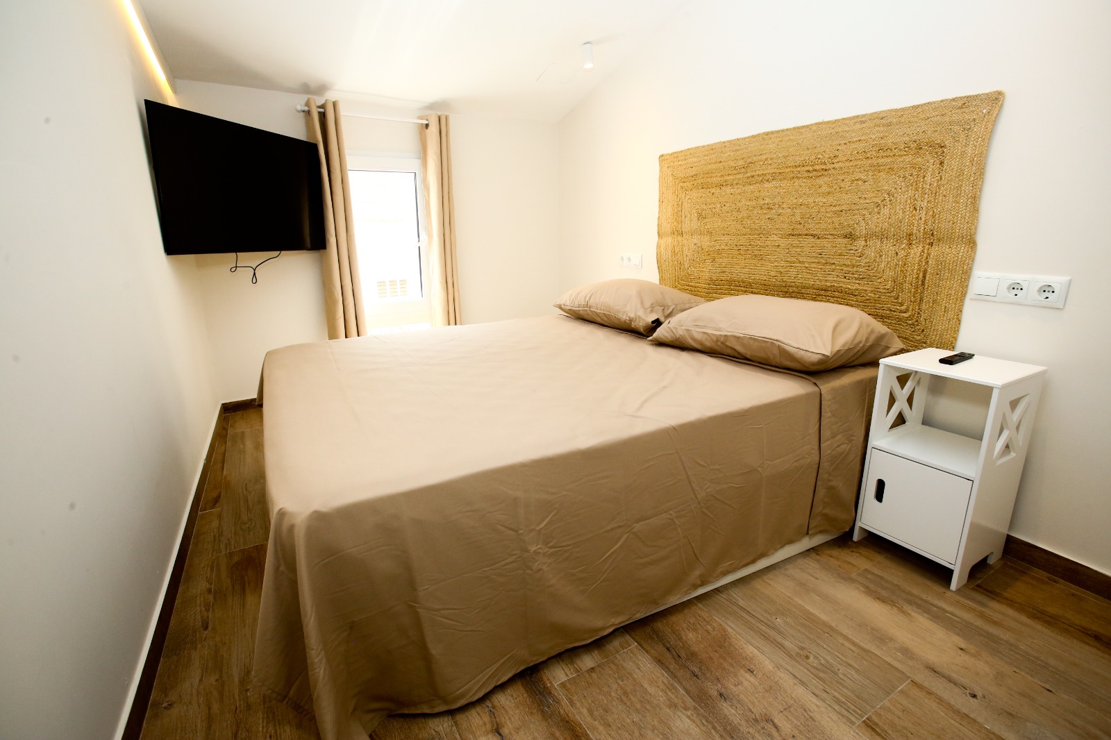 A bedroom with a woven headboard, tan linen, and a window in beige curtains.