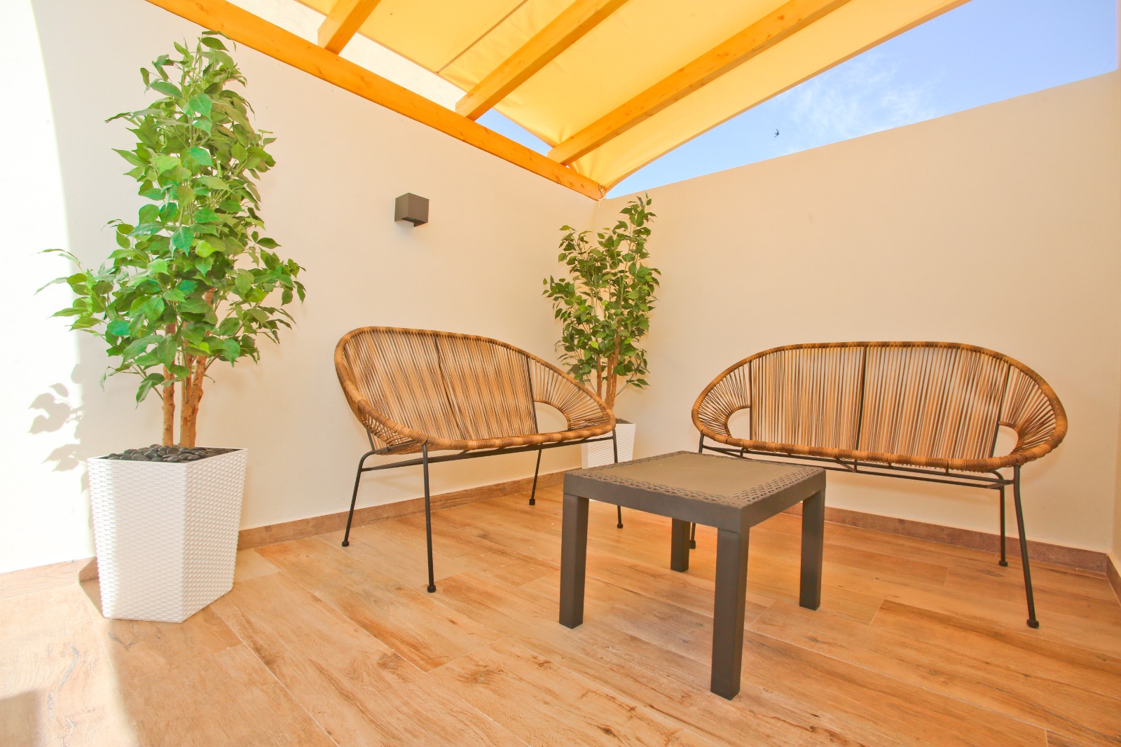 A terrace under a wooden pergola: two woven seats, a small table, white planters.