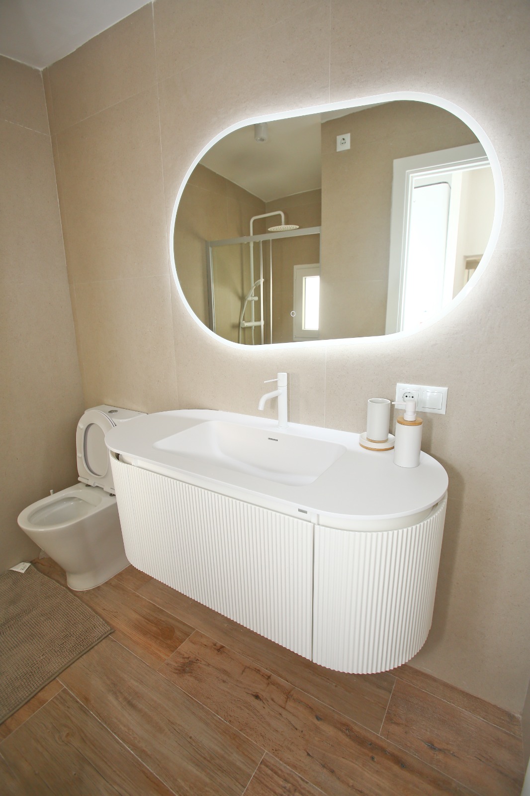 A bathroom with a fluted floating vanity, a backlit capsule mirror, and wood-look floor.