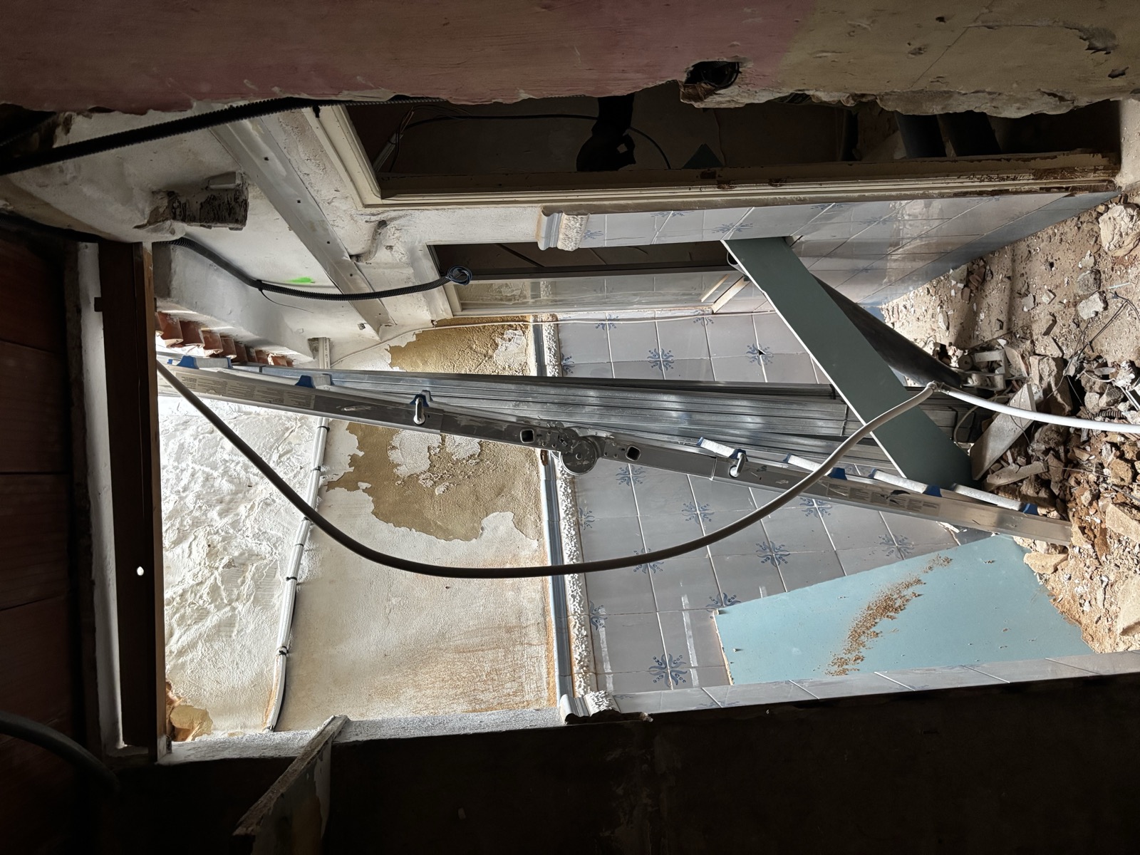 Interior stripped to rubble: old patterned wall tiles, a ladder, hanging wires, daylight through an open roof.